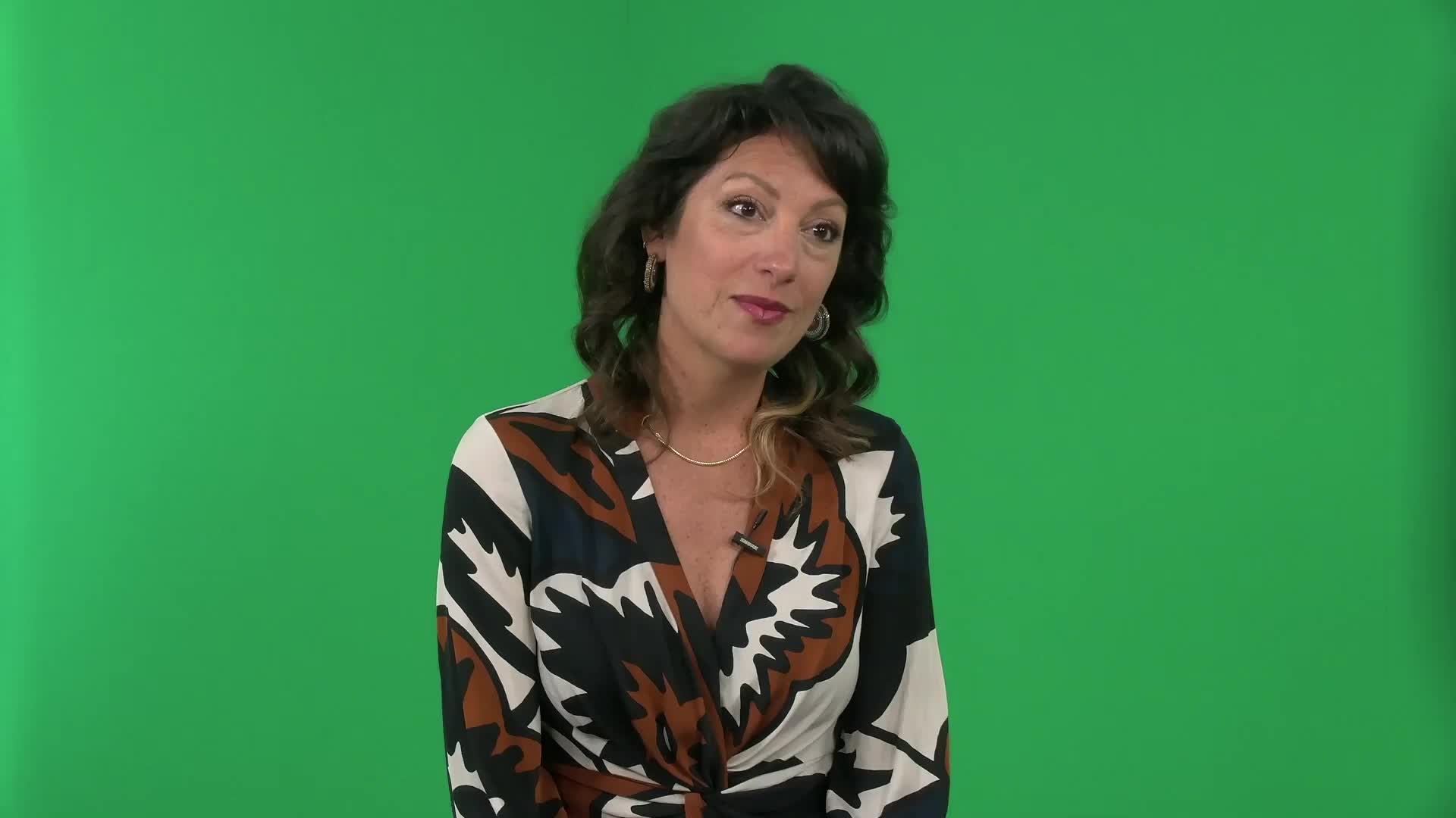 Leah Terranova is a white woman with brown curly hair that is to her shoulders. She is wearing gold metal jewelry, makeup, and a patterned white navy blue and brown blouse. She is smiling and looking to the right of the camera.