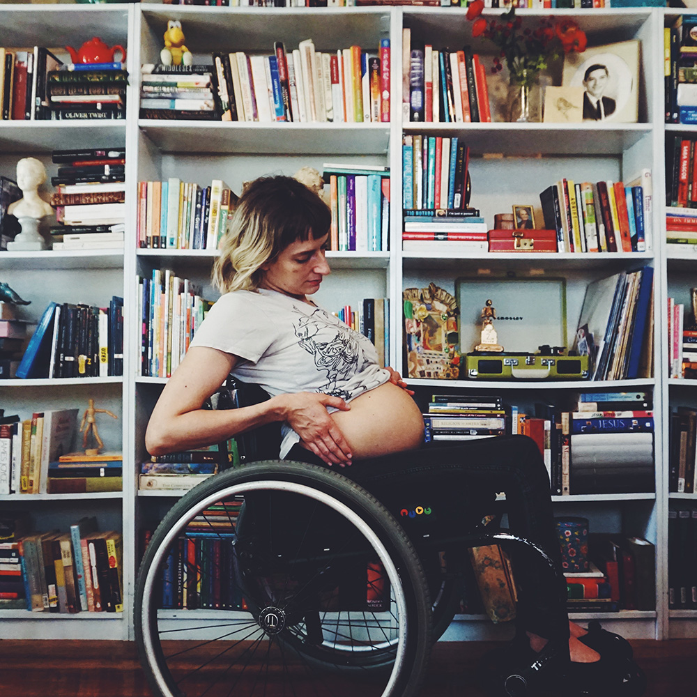The photo captures a profile of Rebekah sitting in her wheelchair in front of a wall of books. She’s lifting up her t-shirt to show a big round belly. She’s leaning back, looking down, her hands on the sides of her belly.