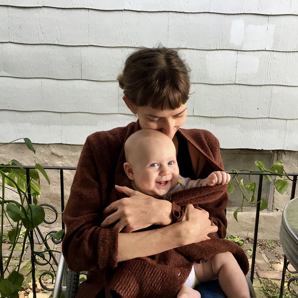 Rebekah sits in her wheelchair with a smiling, bald baby tucked in her massive brown sweater. She rests her lips and cheek against his fuzzy head.