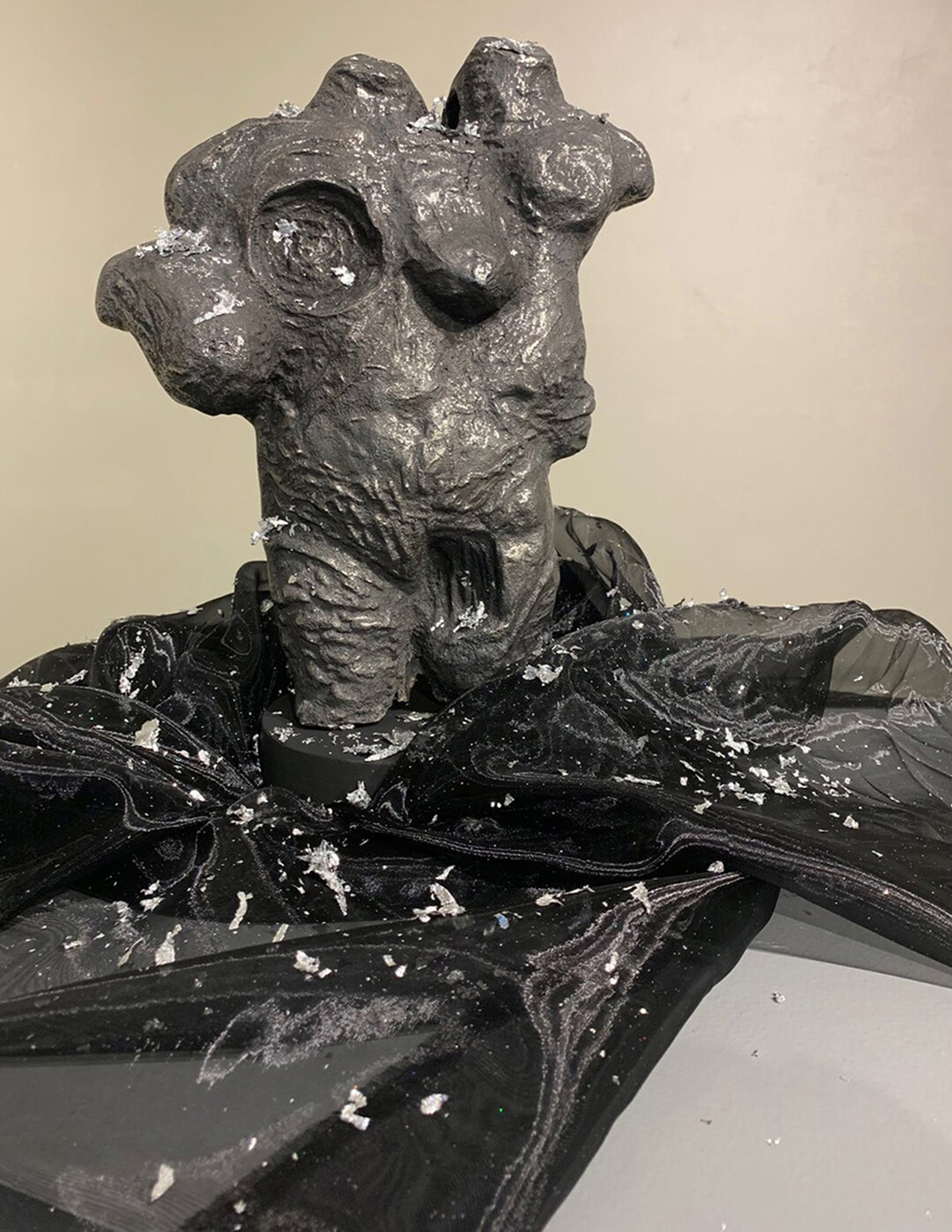 A gray and shiny sculpture stands on top of a black sheer material with silver glitter flakes. The body is morphed to have extra shoulders coming off of the side, and is missing breast tissue in one are and has it added to another portion of the body. There is a gap in the groin area to signify the lack of sex characteristics in that area. There are ridges on the body to signify stretch marks and morphing. The background behind the iron sculpture is a white wall. 
