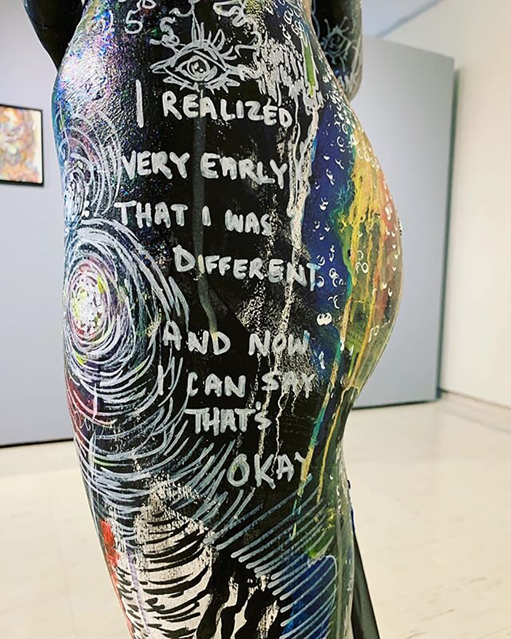 A close up of a mannequin’s lower left side. The mannequin’s side is covered in white, black, blue, and yellow paint with circles, stars, eyeballs, and lines on the side. The words on the side of the mannequin say “I realized very early that I was different and now I can say that’s okay. Behind the mannequin is a light gray and white gallery wall with one painting behind it.