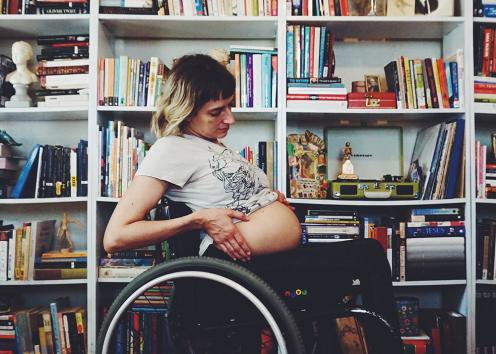 The photo captures a profile of Rebekah sitting in her wheelchair in front of a wall of books. She’s lifting up her t-shirt to show a big round belly. She’s leaning back, looking down, her hands on the sides of her belly. 
