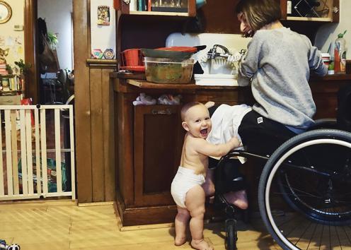 Rebekah sits at the kitchen sink doing dishes from her wheelchair. Her baby stands in his diaper beside her, one arm resting on her wheelchair frame, the other hand holding onto her knee.