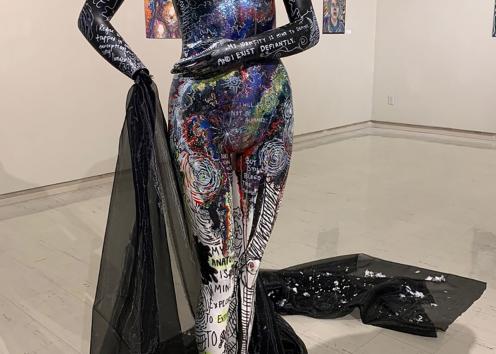 A mannequin stands in a large gallery room. There are paintings on the wall behind the mannequin. The mannequin is black in color mostly with various shades of blues, reds, purples, and greens on it. The legs are white with black and yellow line work. The mannequin is covered in words talking about queer identity. There is a black cloth coming out of the mannequin's wrist that wraps around the mannequin’s feet and trails behind them with silver glitter flakes on the floor. 