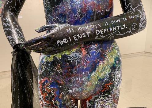A close up of a mannequin’s stomach area and the forearms and left hand. The mannequin’s body is covered in paint varying in blacks, blues, reds, purples, yellows, and greens. The words on the mannequin’s forearm say “My identity is mine to define and I exist defiantly.” The lower half of the mannequin’s body has dots, squiggles, and stars on it and says “I will not be ashamed.” a black sheer fabric exits out of the right wrist of the mannequin. 